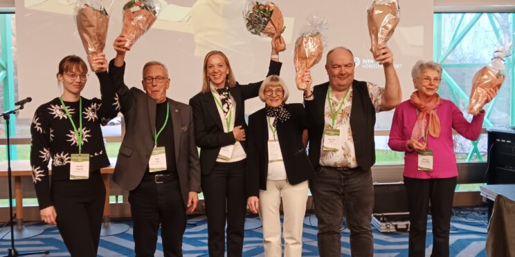 De sex nyvalda styrelsemedlemmarna poserar tillsammans framför kameran. De ler glatt och håller upp blombuketter i luften,