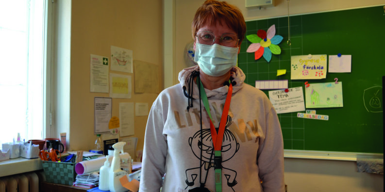 Förskolelärare Maria Eriksson står i sitt klassrum framför sitt skrivbord. Bakom skrivbordet finns en tavla ed olika lappar och papper uppsatta.