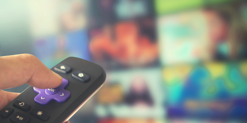 I förgrunden finns en hand som håller i en fjärrkontroll. I bakgrunden finns en påslagen TV.
