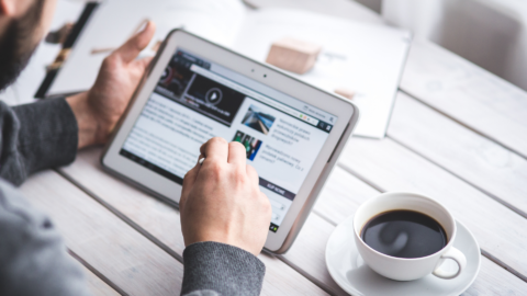 På bilden syns en hand som håller i enen digital surfplatta. Vid sidan om surfplattan finns en kopp med kaffe.