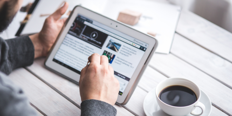 På bilden syns en hand som håller i enen digital surfplatta. Vid sidan om surfplattan finns en kopp med kaffe.