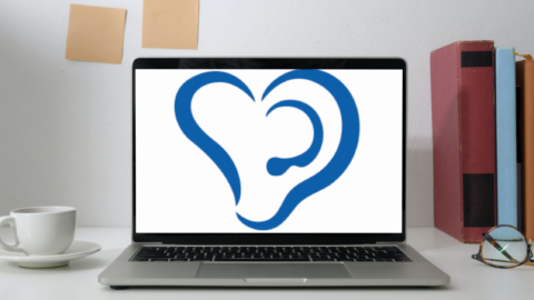 En laptop står på ett skrivbord. På vänster sida finns en kaffekopp och på högra sidan står tre böcker. På bordet ligger ett par glasögon. I laptopens skärm syns Svenska hörselförbundets logo. Logon ser ut som två öron som formar ett hjärta.