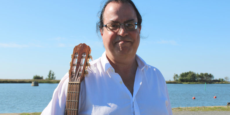 Kalle Teir ler mot kameran och håller en gitarr under högra armen. I bakgrunden syns Bottenhavet och klarblå himmel.