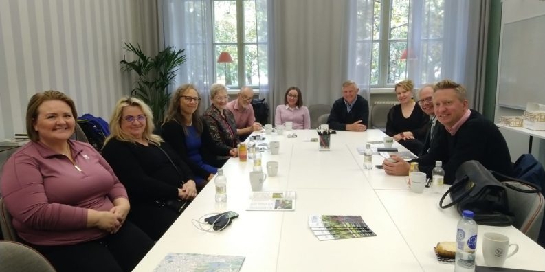 En grupp människor sitter runt ett bord och ler mot kameran. I bakgrunden finns två stora fönster och utanför syns gröna träd och solljus.