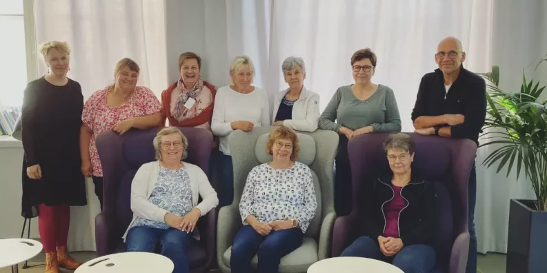 En grupp på tio personer står och sitter vända mot kameran. Rummet är upplyst av solljus som kommer in i rummet genom höga fönster som är täckta av vita gardiner. Personerna ler mot kameran.