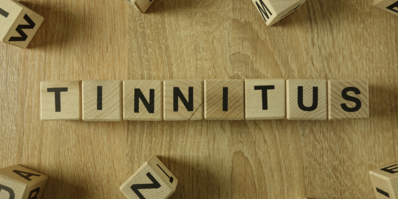 Små träbitar i form av tärningar med bokstäver ligger på ett bord. De är placerade i en rad som formar ordet tinnitus. Runtomkring finns fler träbitar med bokstäver som omger ordet tinnitus.