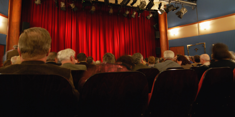 En teatersalong med fullsatt publik. Den röda ridån är nere. I förgrunden ser man åskådares bakhuvuden, bland em finns flera med grått hår.