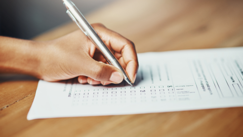 På bilden finns en vänsterhand som håller i en kulspetspenna och fyller i en blankett genom att sätta kryss i rutor.