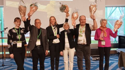 De sex nyvalda styrelsemedlemmarna poserar tillsammans framför kameran. De ler glatt och håller upp blombuketter i luften,